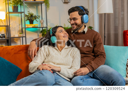 Cheerful happy young Indian couple in wireless headphones dancing on couch at home listening music 120309524