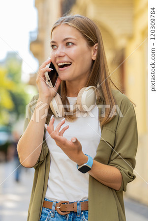 Happy surprised Caucasian young woman in pleasant conversation on smartphone standing on city street 120309784