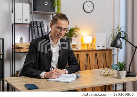 Young businessman looking to camera and writing down on diary in video chat at home office desk Young businessman looking to camera and writing down on diary in video chat at home office desk 120309806