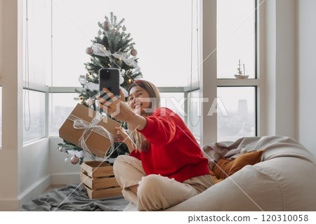 Happy red shirt woman making a video call holiday greeting with her friends and family showing the Christmas tree. Happy red shirt woman making a video call holiday greeting with her friends and family showing the Christmas tree. 120310058