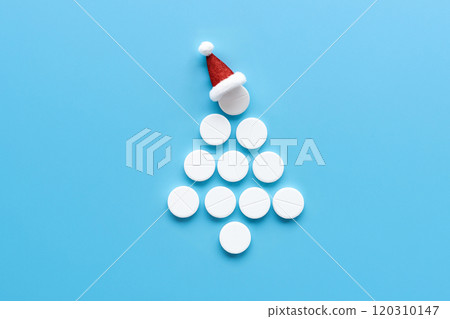 Christmas tree shape made of white pills with Santa hat on top, blue background. Medical holiday concept, flat lay composition, copy space. Unique festive healthcare design 120310147