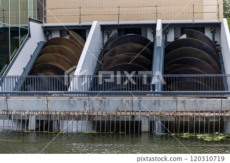 The image shows a set of large Archimedes screws used for water management or energy generation. The screws are housed within a structure beside a canal or river, with a metal railing in front. The image shows a set of large Archimedes screws used for water management or energy generation. The screws are housed within a structure beside a canal or river, with a metal railing in front. 120310719
