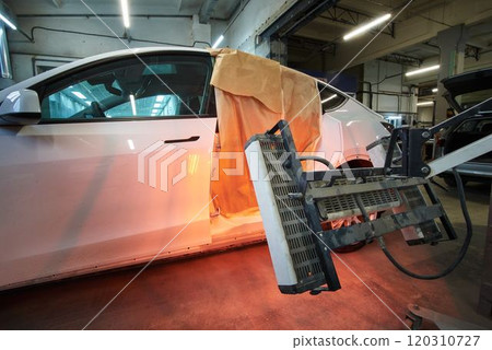 A white car is undergoing body repair with infrared drying equipment. The vehicle's side panel is masked with paper for protection during the heating process in an auto repair shop. A white car is undergoing body repair with infrared drying equipment. The vehicle's side panel is masked with paper for protection during the heating process in an auto repair shop. 120310727