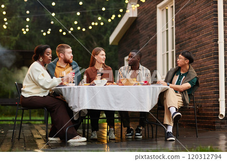 Vertical wide angle shot of multiethnic group of adult friends enjoying dinner together at outdoor patio by brick house copy space 120312794