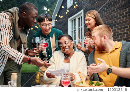 Vibrant shot of multiethnic group of adult friends looking at ultrasound image of Black young woman announcing pregnancy at party outdoors 120312846