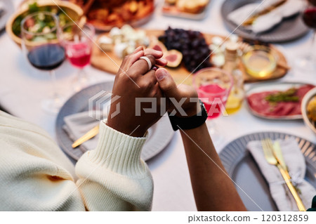 Close up of two people holding hands at dinner table and saying grace during Thanksgiving celebration with friends and family copy space 120312859