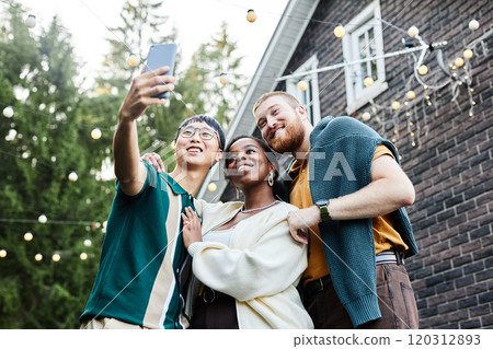 Low angle portrait of multiethnic group of adult friends taking selfie together outdoors in house yard decorated with fairy lights copy space Low angle portrait of multiethnic group of adult friends taking selfie together outdoors in house yard decorated with fairy lights copy space 120312893