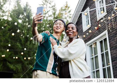 Low angle portrait of smiling adult couple taking selfie together outdoors in house yard with fairy lights copy space 120312894
