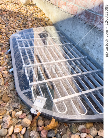 A protective custom plexiglass window well cover with a metal grate surrounded by decorative rocks, placed against a brick wall in a landscaped backyard during the late fall season. 120313089