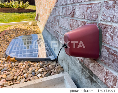 A red exterior vent cover on a brick wall, with a custom plexiglass window well cover surrounded by decorative rocks in a landscaped backyard during the late fall season. A red exterior vent cover on a brick wall, with a custom plexiglass window well cover surrounded by decorative rocks in a landscaped backyard during the late fall season. 120313094