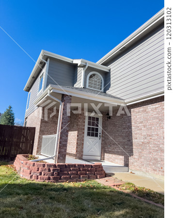 A suburban backyard during late fall, featuring a brick house with a cozy patio and a raised brick planter. The yard includes a green lawn with patches of fallen leaves, a concrete pathway, and a 120313102