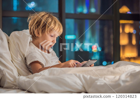 The boy uses the tablet in his bed before going to sleep on a background of a night city. Children and technology concepts The boy uses the tablet in his bed before going to sleep on a background of a night city. Children and technology concepts 120314823