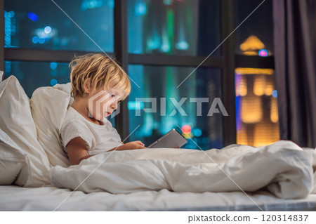 The boy uses the tablet in his bed before going to sleep on a background of a night city. Children and technology concepts The boy uses the tablet in his bed before going to sleep on a background of a night city. Children and technology concepts 120314837