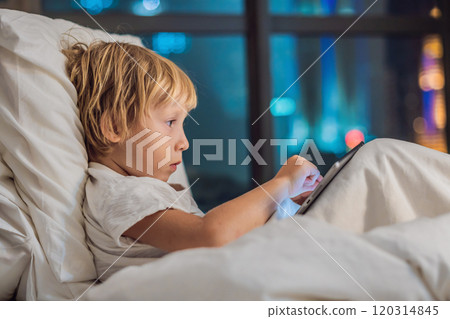 The boy uses the tablet in his bed before going to sleep on a background of a night city. Children and technology concepts 120314845