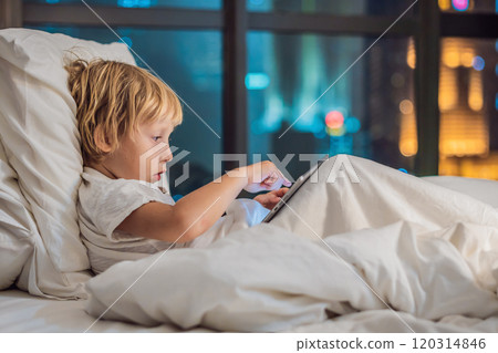 The boy uses the tablet in his bed before going to sleep on a background of a night city. Children and technology concepts The boy uses the tablet in his bed before going to sleep on a background of a night city. Children and technology concepts 120314846