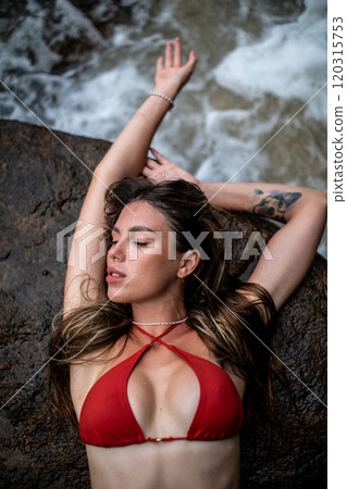 A Young Girl Dressed in a Bright Red Bikini Poses Cheerfully by a Scenic Waterfall 120315753