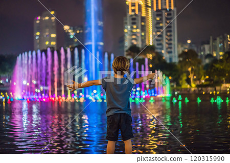 Boy on background of colorful fountain on the lake at night, near by Twin Towers with city on background. Kuala Lumpur, Malaysia Boy on background of colorful fountain on the lake at night, near by Twin Towers with city on background. Kuala Lumpur, Malaysia 120315990