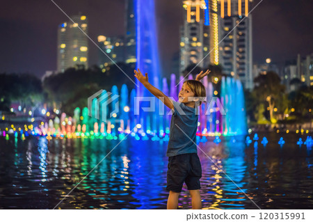 Boy on background of colorful fountain on the lake at night, near by Twin Towers with city on background. Kuala Lumpur, Malaysia Boy on background of colorful fountain on the lake at night, near by Twin Towers with city on background. Kuala Lumpur, Malaysia 120315991