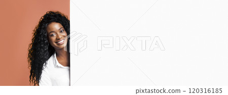 A young black woman with long curly black hair smiles at the camera while peeking from behind a white wall. She is wearing a white button-down shirt and has a peach-colored background behind her. A young black woman with long curly black hair smiles at the camera while peeking from behind a white wall. She is wearing a white button-down shirt and has a peach-colored background behind her. 120316185