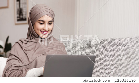 Freelance Concept. Young muslim woman in hijab working on laptop at home, sitting on couch, typing on keyboard and smiling 120316186
