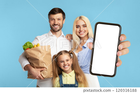 A happy family of three stands in front of a blue background. The mother holds a smartphone with a blank screen towards the camera. The father holds a paper bag full of groceries. A happy family of three stands in front of a blue background. The mother holds a smartphone with a blank screen towards the camera. The father holds a paper bag full of groceries. 120316188