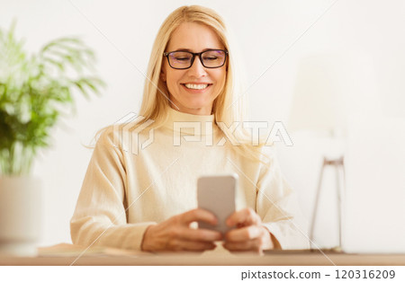 A woman with long blonde hair and glasses smiles while looking at her smartphone. She is wearing a white sweater and is sitting at a table. A woman with long blonde hair and glasses smiles while looking at her smartphone. She is wearing a white sweater and is sitting at a table. 120316209