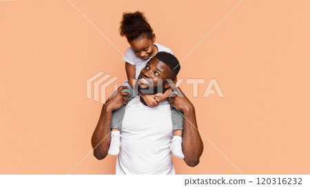 Happy African American father and his daughter posing together against a peach background. The father is smiling and looking to the side while holding his daughter on his shoulders 120316232