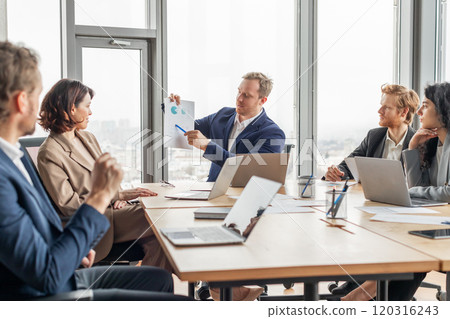 A group of business professionals are gathered around a large conference table in a modern office, engaged in a meeting. One person is presenting charts on a sheet of paper, A group of business professionals are gathered around a large conference table in a modern office, engaged in a meeting. One person is presenting charts on a sheet of paper, 120316243