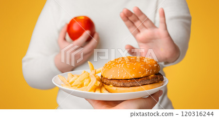 Unrecognizable plus size girl refusing to eat fast junk food, showing stop gesture with hand and choosing apple 120316244