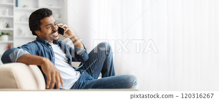 African American man enjoys a moment of relaxation on the couch, engaged in a cheerful phone conversation at his home during the afternoon, surrounded by soft light and a cozy atmosphere. 120316267