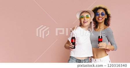 This image features two women in summer hats and sunglasses, smiling and holding soda bottles against a pink background. They are both wearing casual clothing, copy space 120316340