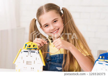 A young girl with long blonde hair, wearing a yellow shirt and denim overalls, is smiling as she carefully assembles a robotic toy. She is holding a wire in her hands, connecting it to the robots body 120316373