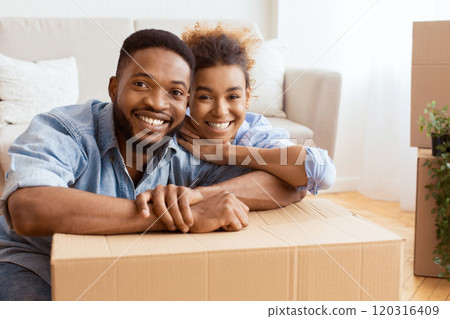 African American Spouses Embracing Among Moving Boxes Smiling At Camera Sitting In New House. Empty Space African American Spouses Embracing Among Moving Boxes Smiling At Camera Sitting In New House. Empty Space 120316409