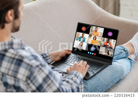 Man sitting on a couch, using a laptop to attend a video conference call. The laptop screen displays several participants in a grid format, indicating that it is an online meeting. Man sitting on a couch, using a laptop to attend a video conference call. The laptop screen displays several participants in a grid format, indicating that it is an online meeting. 120316415