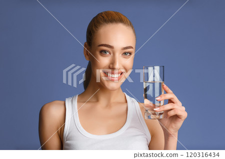 This image shows a young woman with a bright smile holding a glass of water in her right hand. She is wearing a white tank top. The background is a solid blue color. 120316434