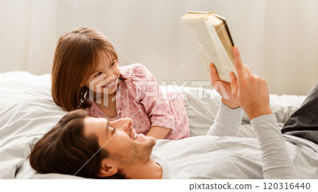 Young man reading book to his little smiling daughter, laying on bed together Young man reading book to his little smiling daughter, laying on bed together 120316440