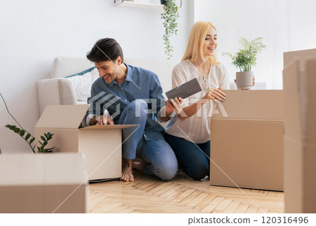 A young couple unpacks boxes in their new home, surrounded by other boxes. They are smiling and look excited about their new space. 120316496