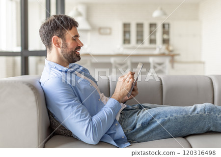 A man in a blue shirt and jeans sits comfortably on a gray couch, scrolling through a tablet. He leans back and smiles, looking relaxed and content. The room is bright and modern A man in a blue shirt and jeans sits comfortably on a gray couch, scrolling through a tablet. He leans back and smiles, looking relaxed and content. The room is bright and modern 120316498