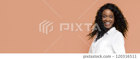 A smiling Black woman with long curly hair is wearing a white button-up shirt against a peach-colored background. She looks to the side with a genuine smile. A smiling Black woman with long curly hair is wearing a white button-up shirt against a peach-colored background. She looks to the side with a genuine smile. 120316511