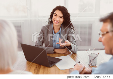 Offering Tours. Smiling mexican travel agent working with happy mature clients in office, gesturing, using laptop Offering Tours. Smiling mexican travel agent working with happy mature clients in office, gesturing, using laptop 120316533