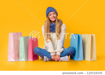 Shop as you can. Carefree blonde girl in winter clothes sitting on floor among shopping bags, pink studio background, copy space Shop as you can. Carefree blonde girl in winter clothes sitting on floor among shopping bags, pink studio background, copy space 120316545