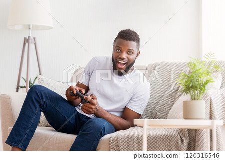 A young black man sits on a couch in his living room, excitedly playing video games using a black controller. He is wearing a white t-shirt and jeans, and there is a plant on a side table behind him. 120316546