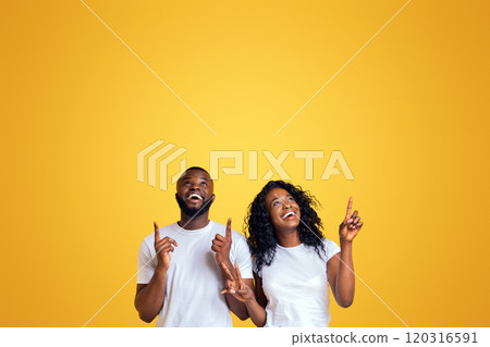 Smiling Black Couple Is Pointing Fingers Upwards At Free Space, yellow studio background Smiling Black Couple Is Pointing Fingers Upwards At Free Space, yellow studio background 120316591