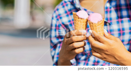 A close-up image of a person holding two ice cream cones with pink ice cream in them. The person is wearing a plaid shirt and is outdoors on a sunny day. The background is blurred and out of focus. A close-up image of a person holding two ice cream cones with pink ice cream in them. The person is wearing a plaid shirt and is outdoors on a sunny day. The background is blurred and out of focus. 120316595