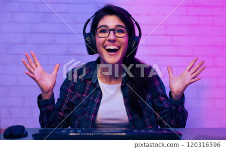 This is a photo of a woman wearing headphones and a plaid shirt, seated in front of a computer with her arms raised. She is smiling broadly and looks excited. She is likely playing a video game. This is a photo of a woman wearing headphones and a plaid shirt, seated in front of a computer with her arms raised. She is smiling broadly and looks excited. She is likely playing a video game. 120316596