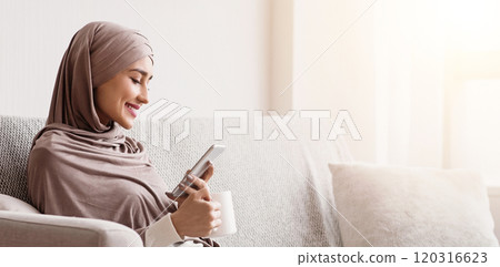 Weekend Relax. Muslim Girl In Hijab Resting At Home With Smartphone And Coffee, Sitting On Sofa In Living Room, Free Space Weekend Relax. Muslim Girl In Hijab Resting At Home With Smartphone And Coffee, Sitting On Sofa In Living Room, Free Space 120316623