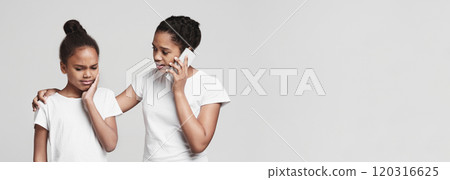 Little african girl suffering from tooth pain, her teen sister calling ambulance, grey studio background 120316625