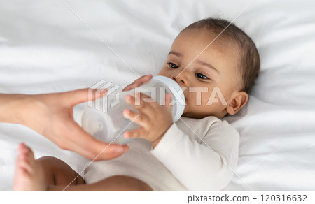 Childcare And Nutrition Concept. Portrait of cute little African American baby drinking milk from bottle, woman helping and holding, kid wearing bodysuit lying on the white blanket at home Childcare And Nutrition Concept. Portrait of cute little African American baby drinking milk from bottle, woman helping and holding, kid wearing bodysuit lying on the white blanket at home 120316632
