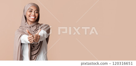 Positive afro muslim girl in hijab gesturing thumbs up and smiling at camera while posing over yellow background in studio, empty space 120316651