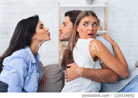 Love Triangle And Jealousy. Cheating Boyfriend Kissing A Girl While Hugging His Girlfriend Sitting Together On Couch Indoor. 120316692